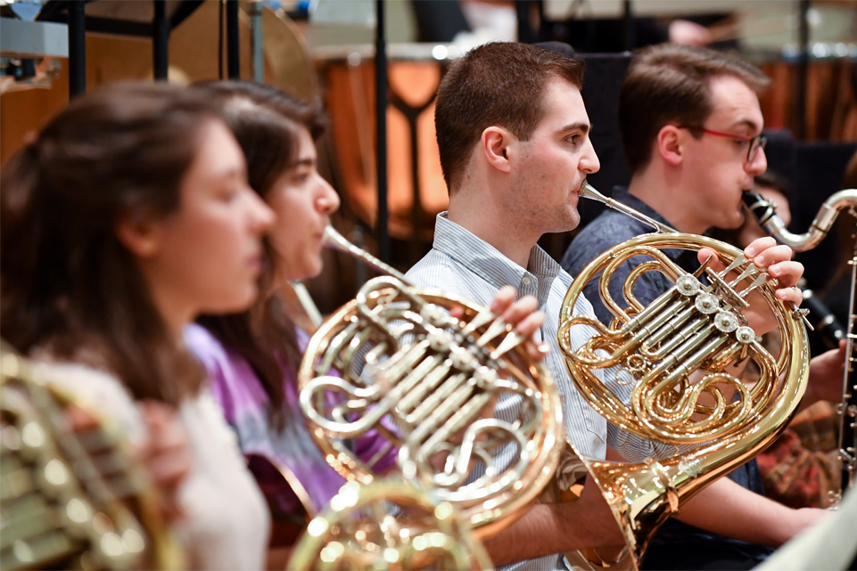 A group of students, performing and holding a French horn in a orchestra rehearsal.